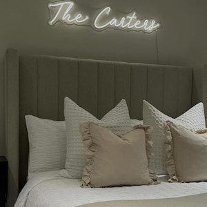 K&ouml;nnte beinhalten: Ein Schlafzimmer mit einem Bett mit grauem Kopfteil und wei&szlig;er Bettw&auml;sche. &Uuml;ber dem Bett steht in einem Neon-Schild "The Carters" in Schreibschrift. Dekorative Kissen in Wei&szlig; und Beige verleihen dem Bett Textur und Farbe.