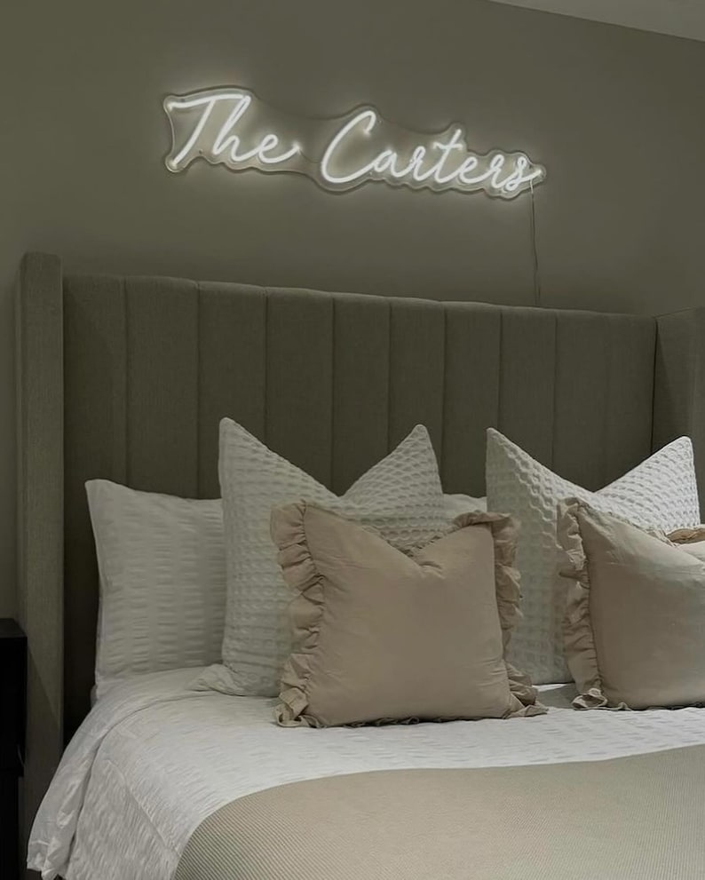 K&ouml;nnte beinhalten: Ein Schlafzimmer mit einem Bett mit grauem Kopfteil und wei&szlig;er Bettw&auml;sche. &Uuml;ber dem Bett steht in einem Neon-Schild "The Carters" in Schreibschrift. Dekorative Kissen in Wei&szlig; und Beige verleihen dem Bett Textur und Farbe.