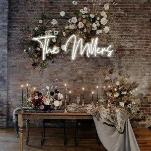 K&ouml;nnte beinhalten: Hochzeitsdekoration mit einem rustikalen Holztisch, Blumengestecken und Kerzen. Ein Neonschild mit der Aufschrift "The Millers" vor einer Backsteinmauer. Der Tisch ist mit einem grauen Tuch drapiert.