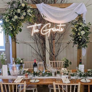 K&ouml;nnte beinhalten: Ein Hochzeitsempfangstisch mit Blumenschmuck, Gl&auml;sern und einem Neonschild mit der Aufschrift "The Grants". Der Holztisch ist mit Gr&uuml;n geschm&uuml;ckt. Eine Weinflasche und eine Wasserkaraffe sind sichtbar.