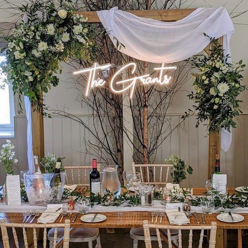 K&ouml;nnte beinhalten: Ein Hochzeitsempfangstisch mit Blumenschmuck, Gl&auml;sern und einem Neonschild mit der Aufschrift "The Grants". Der Holztisch ist mit Gr&uuml;n geschm&uuml;ckt. Eine Weinflasche und eine Wasserkaraffe sind sichtbar.
