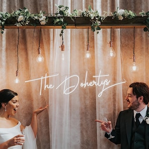 K&ouml;nnte beinhalten: Hochzeitsfeier mit einem Neon-Schild mit der Aufschrift "The Dohertys". Das Schild leuchtet wei&szlig; und h&auml;ngt vor einem transparenten Vorhang. Die Szene umfasst h&auml;ngende Gl&uuml;hbirnen und Blumenschmuck.