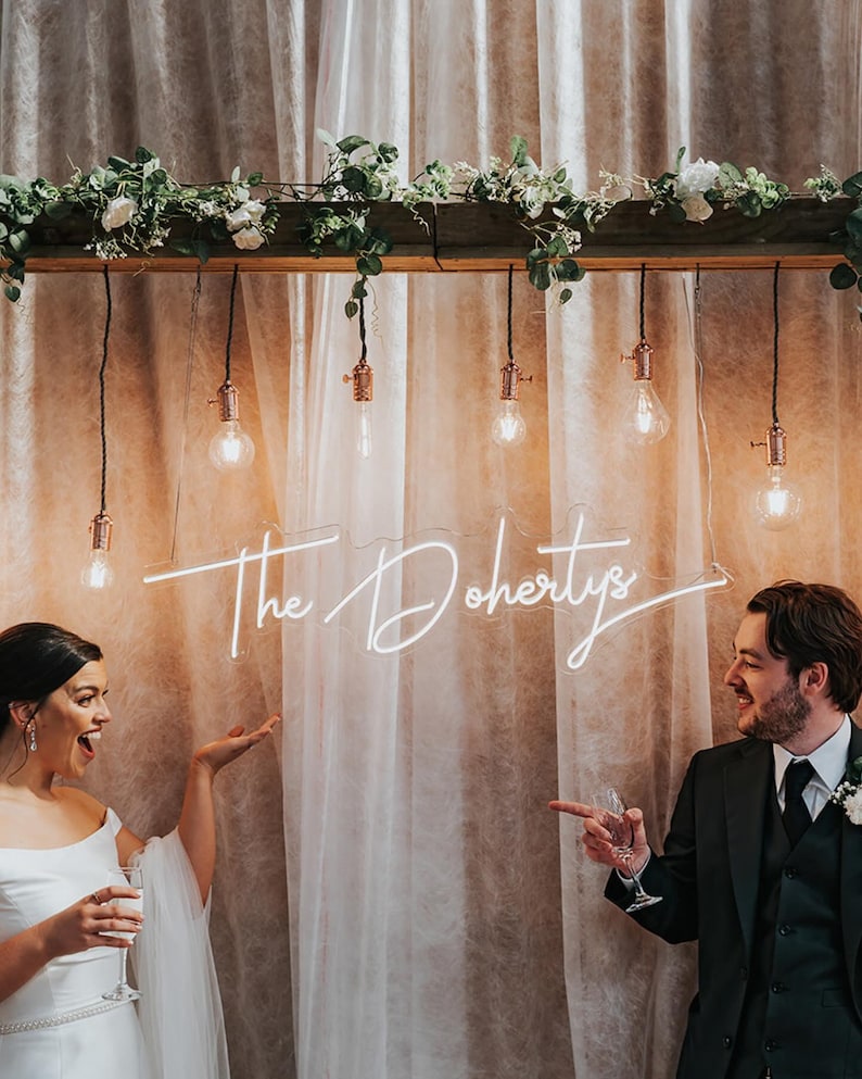 K&ouml;nnte beinhalten: Hochzeitsfeier mit einem Neon-Schild mit der Aufschrift "The Dohertys". Das Schild leuchtet wei&szlig; und h&auml;ngt vor einem transparenten Vorhang. Die Szene umfasst h&auml;ngende Gl&uuml;hbirnen und Blumenschmuck.