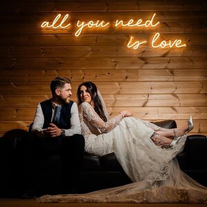 K&ouml;nnte beinhalten: Ein Paar sitzt auf einem schwarzen Sofa vor einer Holzwand. Die Frau tr&auml;gt ein wei&szlig;es Spitzenhochzeitskleid und der Mann eine Weste und Krawatte. Ein Neon-Schild &uuml;ber ihnen lautet "all you need is love."