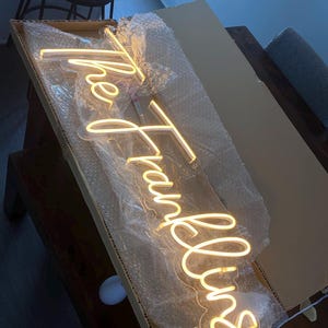 K&ouml;nnte beinhalten: Ein warmwei&szlig;es Neon-Schild in einem Karton mit der Aufschrift "the Franklins" in einer Schreibschrift. Das Schild ist in Luftpolsterfolie eingeschlossen. Die Box befindet sich auf einer dunklen Holzoberfl&auml;che.