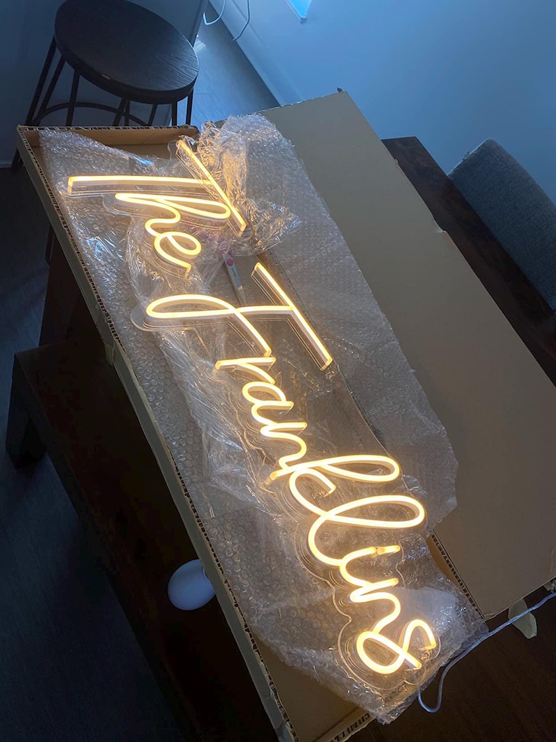 K&ouml;nnte beinhalten: Ein warmwei&szlig;es Neon-Schild in einem Karton mit der Aufschrift "the Franklins" in einer Schreibschrift. Das Schild ist in Luftpolsterfolie eingeschlossen. Die Box befindet sich auf einer dunklen Holzoberfl&auml;che.