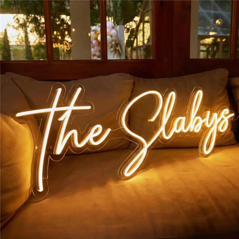 K&ouml;nnte beinhalten: Ein Neonschild mit der Aufschrift "The Slabys" in Schreibschrift. Das Schild leuchtet mit warmwei&szlig;em Licht und ist auf einer transparenten R&uuml;ckwand montiert. Das Schild befindet sich auf einem beigen Sofa.