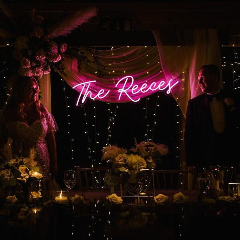 K&ouml;nnte beinhalten: Eine Hochzeitsfeier mit einem pinkfarbenen Neon-Schild mit der Aufschrift "The Reeces". Der Tisch ist mit Blumen und Kerzen geschm&uuml;ckt. Braut und Br&auml;utigam stehen auf beiden Seiten des Tisches.