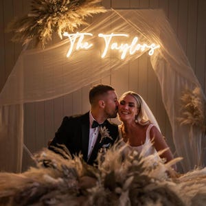 K&ouml;nnte beinhalten: Hochzeitsszene mit einem Paar unter einem Neon-Schild "The Taylors". Die Braut tr&auml;gt ein wei&szlig;es Kleid und Schleier, der Br&auml;utigam einen schwarzen Smoking. Pampasgras schm&uuml;ckt die Szene und schafft eine romantische Atmosph&auml;re.