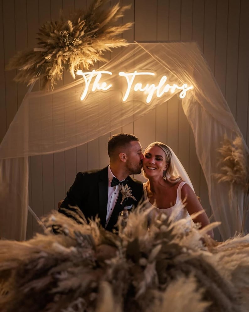 K&ouml;nnte beinhalten: Hochzeitsszene mit einem Paar unter einem Neon-Schild "The Taylors". Die Braut tr&auml;gt ein wei&szlig;es Kleid und Schleier, der Br&auml;utigam einen schwarzen Smoking. Pampasgras schm&uuml;ckt die Szene und schafft eine romantische Atmosph&auml;re.
