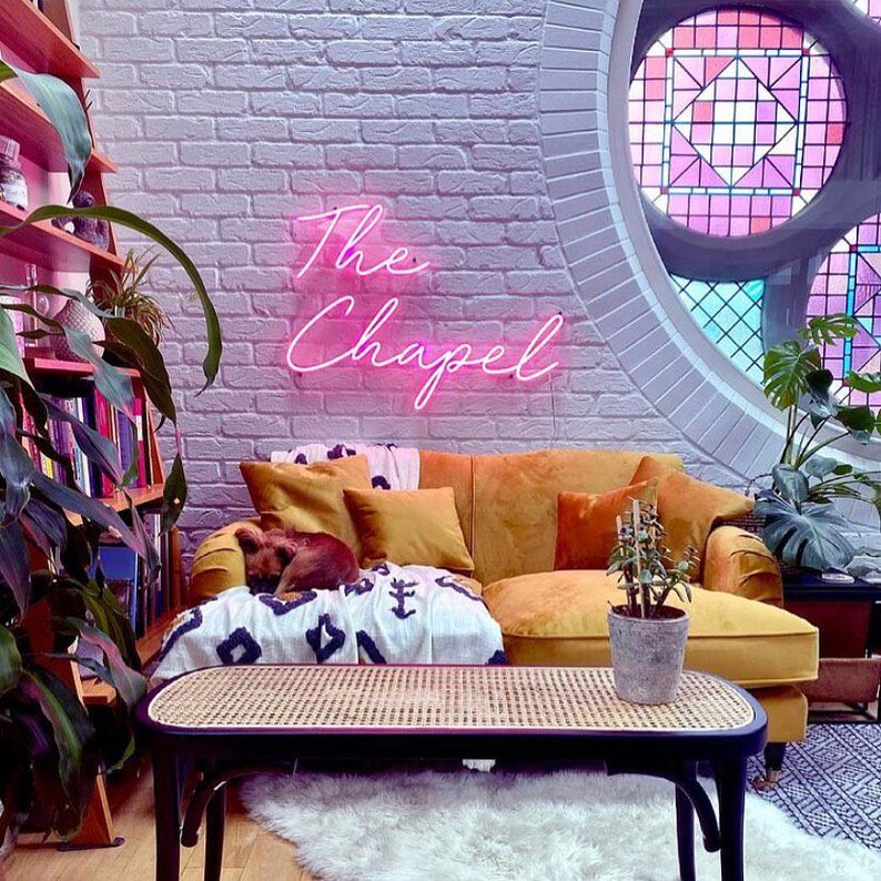 K&ouml;nnte beinhalten: Ein gem&uuml;tliches Wohnzimmer mit einem senffarbenen Sofa, Kissen und einer gemusterten Decke. Ein Neonschild mit der Aufschrift "The Chapel" h&auml;ngt an einer Backsteinmauer. Ein gewebter Couchtisch und ein Buntglasfenster erg&auml;nzen die Einrichtung.