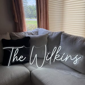 K&ouml;nnte beinhalten: Ein wei&szlig;es Neon-Schild mit dem Schriftzug "The Wilkins" in Schreibschrift. Das Schild liegt auf einem hellgrauen Ecksofa mit einem schwarzen Kissen. Der Hintergrund zeigt ein Fenster mit Blick auf B&auml;ume und einen braunen Vorhang.