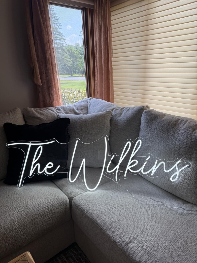 K&ouml;nnte beinhalten: Ein wei&szlig;es Neon-Schild mit dem Schriftzug "The Wilkins" in Schreibschrift. Das Schild liegt auf einem hellgrauen Ecksofa mit einem schwarzen Kissen. Der Hintergrund zeigt ein Fenster mit Blick auf B&auml;ume und einen braunen Vorhang.