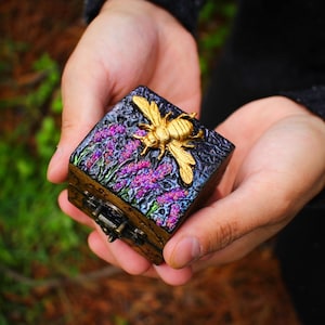 Peut inclure: Une petite boîte carrée avec un couvercle à charnière, décorée d'un motif floral violet et noir et d'une abeille dorée. La boîte a un loquet en bronze.