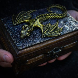 May include: A wooden box with a gold dragon sculpture on the lid. The dragon has a detailed design and is lying on its side with its wings spread out. The box has a dark wood finish and a metal latch.