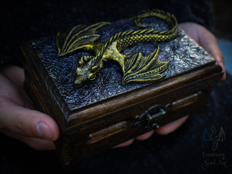 May include: A wooden box with a gold dragon sculpture on the lid. The dragon has a detailed design and is lying on its side with its wings spread out. The box has a dark wood finish and a metal latch.
