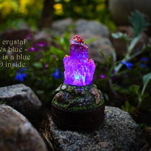 May include: A purple crystal glows with a blue LED light inside. The crystal is surrounded by small rocks and moss. The text "The crystal glows blue - there is a blue LED inside" is visible in the image.