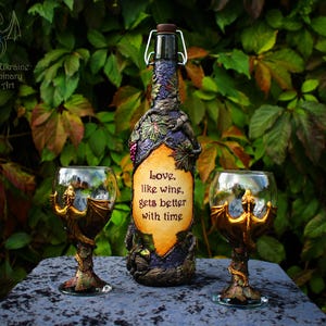 Pode incluir: Uma garrafa de vinho decorativa e duas taças de vinho a condizer. A garrafa é adornada com detalhes esculpidos e um rótulo que diz "Love, like wine, gets better with time". As taças apresentam hastes em forma de dragão.