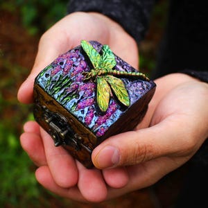 May include: A small square wooden box with a hinged metal latch. The lid features a textured purple and blue surface decorated with a raised, iridescent green dragonfly sculpture and purple floral accents. The box has a dark, weathered finish.