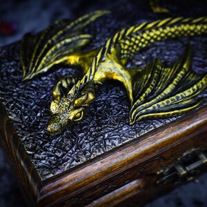 May include: A wooden box with a golden dragon sculpture on the lid. The dragon has detailed wings and scales, and its eyes are open. The box has a dark brown finish and a metal latch.