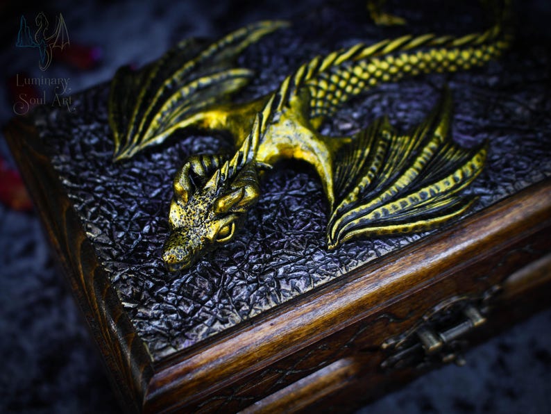 May include: A wooden box with a golden dragon sculpture on the lid. The dragon has detailed wings and scales, and its eyes are open. The box has a dark brown finish and a metal latch.
