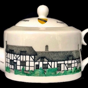 Könnte beinhalten: Weiße Porzellan-Teekanne mit einer Schwarzweiß-Illustration eines Hauses mit Strohdach. Die Teekanne hat ein gelbes und schwarzes Wappen auf dem Deckel und den Text "Home of the Blackamoor's Wife - Built 1516" an der Seite.