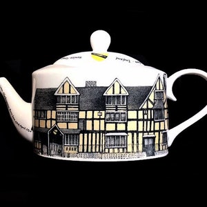 Può includere: Teiera in ceramica bianca con un'illustrazione in nero e giallo di una casa in stile Tudor. La casa è decorata con travi in legno nero e bianco e accenti gialli. La teiera ha un manico e un beccuccio neri.