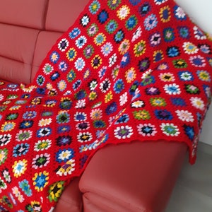 Può includere: Una coperta di uncinetto rossa con un motivo a quadrati di nonna multicolore. La coperta è drappeggiata su un divano in pelle rossa.