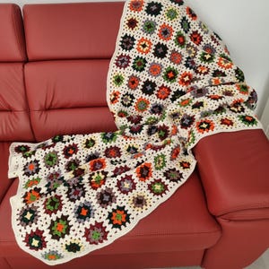 May include: A crocheted throw blanket with a granny square pattern in shades of white, beige, brown, green, orange, and red. The blanket is draped over a red leather sofa.