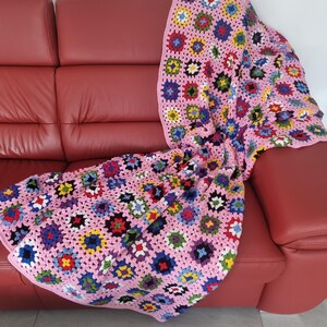 Peut inclure: Une couverture en crochet rose avec des carrés de grand-mère multicolores. La couverture est posée sur un canapé en cuir rouge.