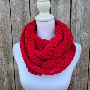 May include: A vibrant red, plush infinity scarf displayed on a white mannequin. The scarf is made of a soft, textured material, and is wrapped around the mannequin's neck. The background is a weathered wooden fence.