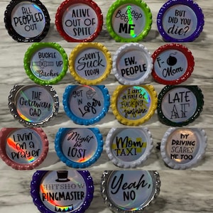 May include: A collection of colorful bottle caps with various phrases and sayings in black text. The caps have different colored edges, including black, red, green, yellow, blue, and white. Some phrases include "All Peopled Out," "Alive Out of Spite," and "But Did You Die?"
