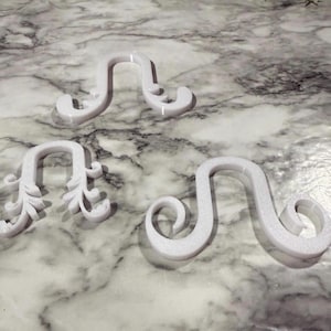 May include: Three white, U-shaped hooks with decorative scrollwork ends. Each hook features a distinct curved design, with one displaying leaf-like patterns along the sides, while the other two exhibit smooth, rounded, and curled terminal ends.
