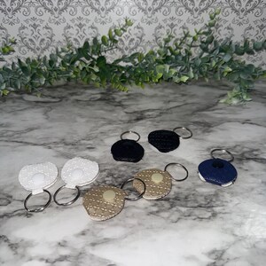 May include: A collection of keychains in various colors and textures. The keychains feature round or oval shapes with a metal ring. Colors include white, black, navy blue, and tan. Some have textured surfaces, while others have a smooth appearance.