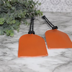 May include: Two orange leather keychains with black cords. The keychains are shaped like a rounded rectangle.