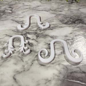 May include: Three white decorative wall hooks with a textured finish. The hooks have a curved design with decorative flourishes. The hooks are arranged on a marble-patterned surface, with greenery in the background.