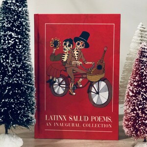 Peut inclure: Un livre reli&eacute; rouge avec le titre "Latinx Salud Poems, An Inaugural Collection" et une illustration de deux squelettes &agrave; v&eacute;lo. Les squelettes portent des chapeaux et l'un d'eux tient une guitare.