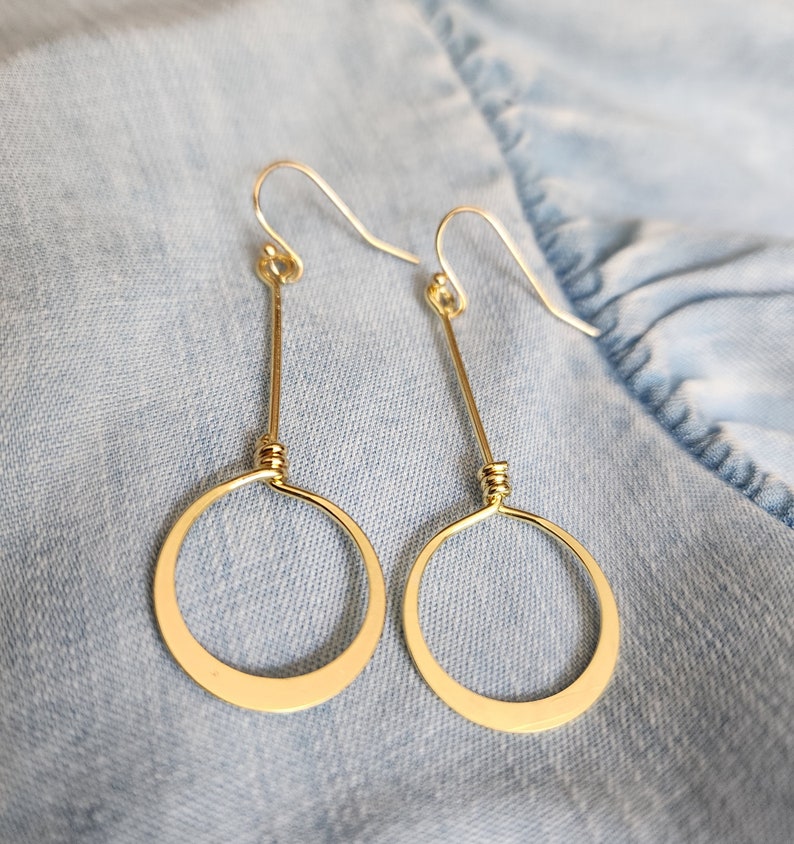 May include: A pair of gold dangle earrings with a simple circle design. The earrings are made of wire and have a delicate, minimalist look.