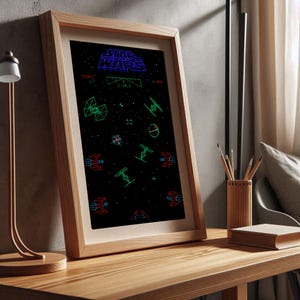 May include: A framed print of the classic Star Wars arcade game. The print features a black background with green, blue, and red pixelated graphics of spaceships and the text "STAR WARS".