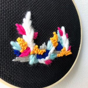 Peut inclure: Gros plan d'un cercle de broderie noir avec un motif floral coloré brodé dessus. Le motif est composé de fils blancs, roses, bleus, jaunes et argentés.