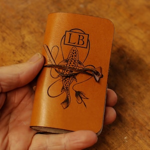 May include: A small, tan leather notebook with a fish illustration and the initials "LB" in a rectangle. The notebook is secured with a brown cord. The background is a wooden surface.