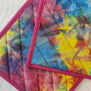 May include: Two quilted hot pads with a tie-dye pattern in shades of pink, yellow, blue, and purple. The hot pads have a pink border and are quilted with a diamond pattern.