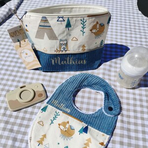 May include: A blue and white baby bib with a fox and trees design. The bib has a blue corduroy trim and the name "Mathias" embroidered on it. A white and blue toiletry bag with a fox and trees design is also pictured. The bag has the name "Mathias" embroidered on it. A wooden camera toy and a clear baby bottle are also pictured.