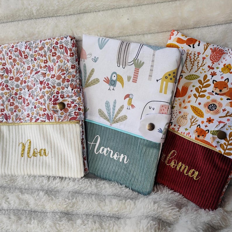 May include: Three personalized fabric baby books with different patterns. The first book has a floral pattern with the name "Mia" embroidered on the front. The second book has a jungle animal pattern with the name "Aaron" embroidered on the front. The third book has a fox pattern with the name "Loma" embroidered on the front.