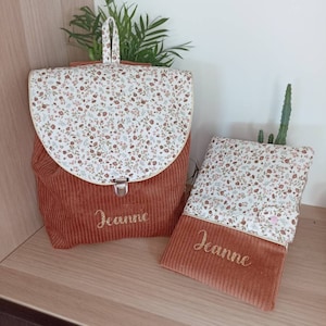 May include: A set of two brown corduroy bags with a floral print lining. The larger bag is a backpack with a zipper closure and the smaller bag is a flat pouch. Both bags are personalized with the name "Jeanne" embroidered in gold thread.