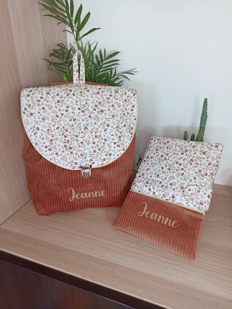 May include: A set of two brown corduroy bags with a floral print lining. The larger bag is a backpack with a zipper closure and the smaller bag is a flat pouch. Both bags are personalized with the name "Jeanne" embroidered in gold thread.