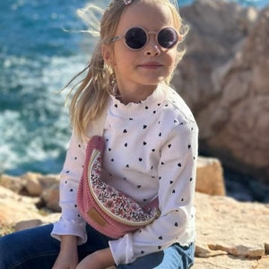Può includere: Una bambina che indossa una camicia bianca a maniche lunghe con cuori neri stampati, occhiali da sole rotondi rosa e una marsupio in velluto rosa con stampa floreale.