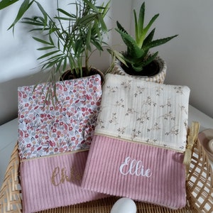 May include: Two pink fabric pouches with gold zippers and embroidered names. The pouch on the left has the name "Chloe" embroidered in gold glitter. The pouch on the right has the name "Ellie" embroidered in white. Both pouches have a floral pattern on the top half and a pink corduroy texture on the bottom half.