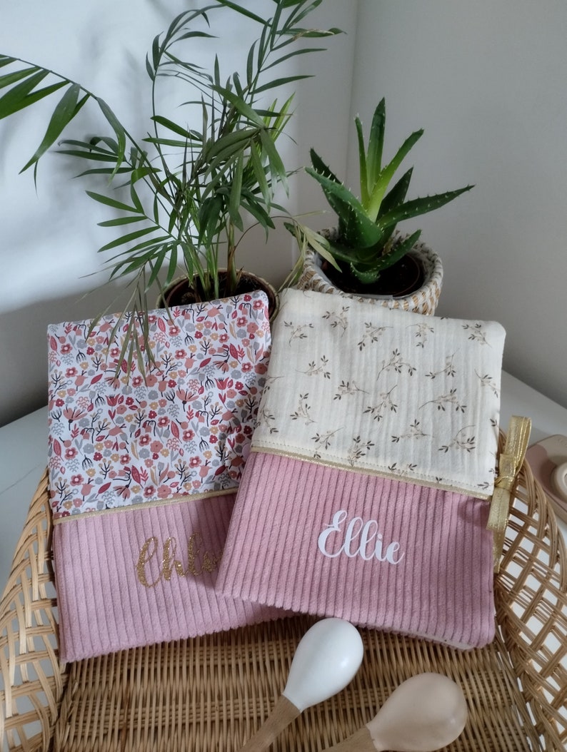 May include: Two pink fabric pouches with gold zippers and embroidered names. The pouch on the left has the name "Chloe" embroidered in gold glitter. The pouch on the right has the name "Ellie" embroidered in white. Both pouches have a floral pattern on the top half and a pink corduroy texture on the bottom half.