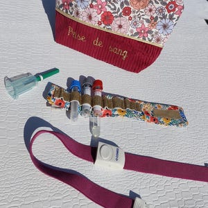 May include: A floral print cosmetic bag with the text "Prise de sang" in gold embroidery. Also shown are a tourniquet, blood collection tubes, and a needle holder, all for medical use.